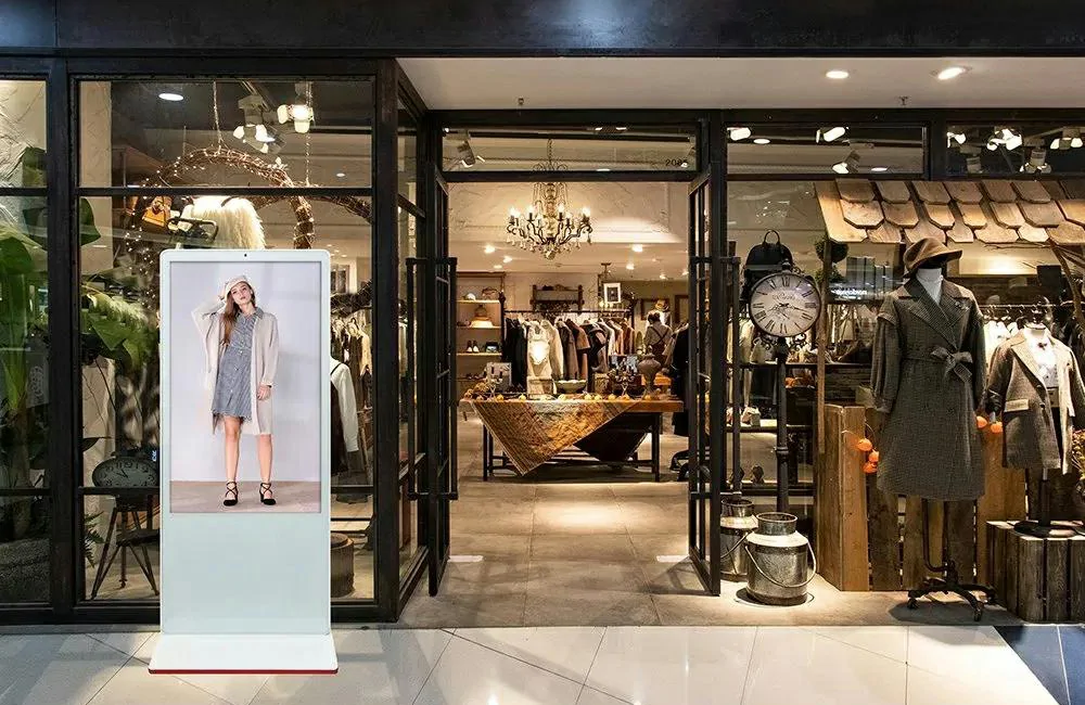 White vertical LCD poster display stand placed at clothing store entrance to promote new women apparel collection